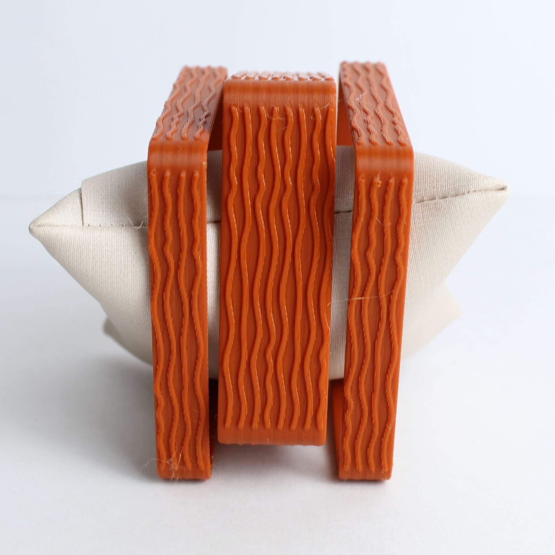 Pulseiras terracota empilhadas em PLA com textura ondulada, exibidas em almofada de apoio