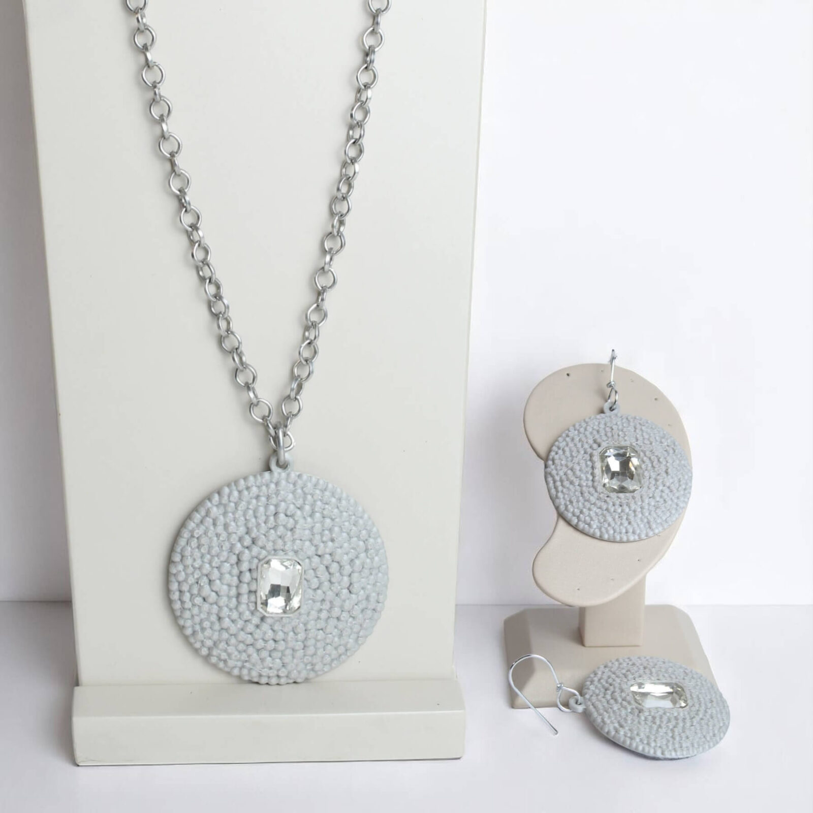 Conjunto composto por colar e brinco em PLA prateado com textura e pedra transparente, expostos em suporte